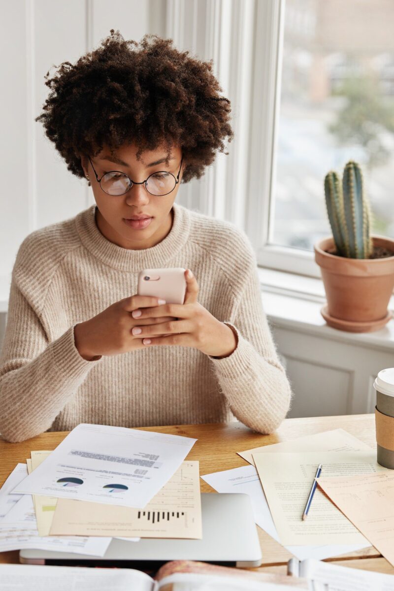 busy-businesswoman-with-afro-hairstyle-works-with-documentation-checks-bank-account-via-cell-phone.jpg